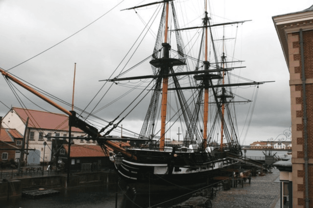 Historic sailing ship docked at a harbor with overcast skies and nearby buildings. - Home Instead