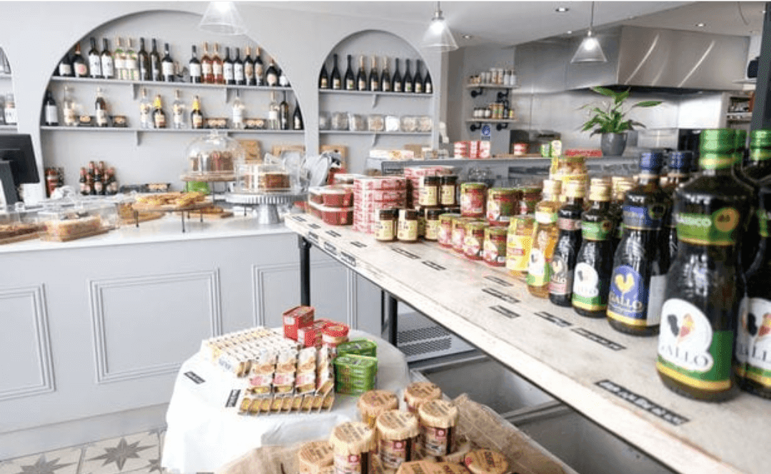 Interior of a gourmet grocery store with shelves displaying various wines, preserves, oils, and breads in a bright setting. - Home Instead