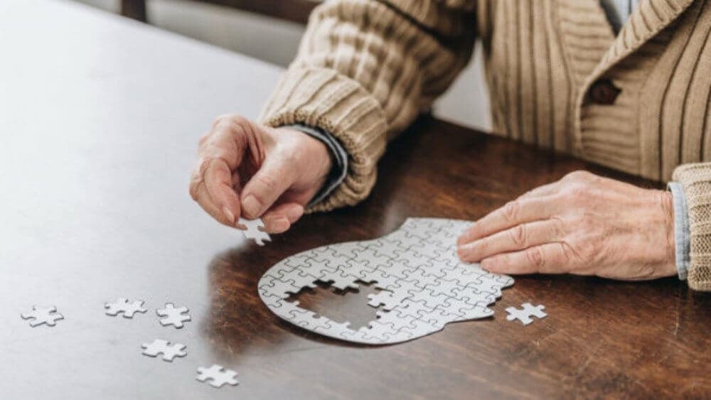 Elderly person assembling a white jigsaw puzzle in the shape of a head on a wooden table. - Home Instead