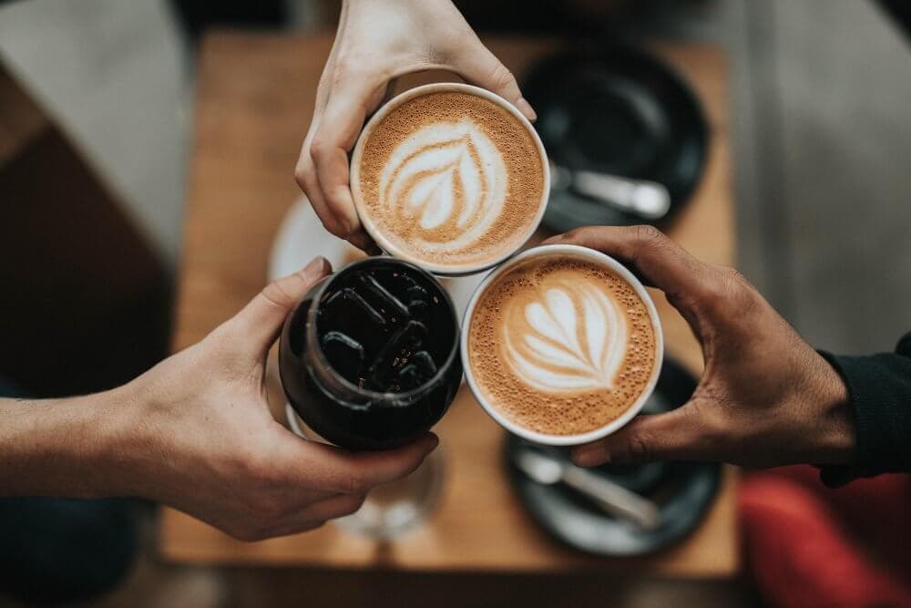 Three hands holding latte art coffee cups and a glass of iced coffee, all toasting over a table with black saucers. - Home Instead