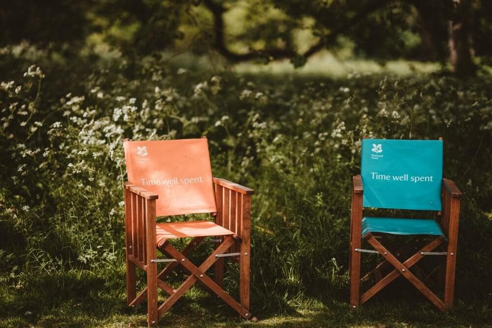 Two wooden director’s chairs, one orange and one teal, with "Time well spent" printed on their backs, in a lush garden. - Home Instead