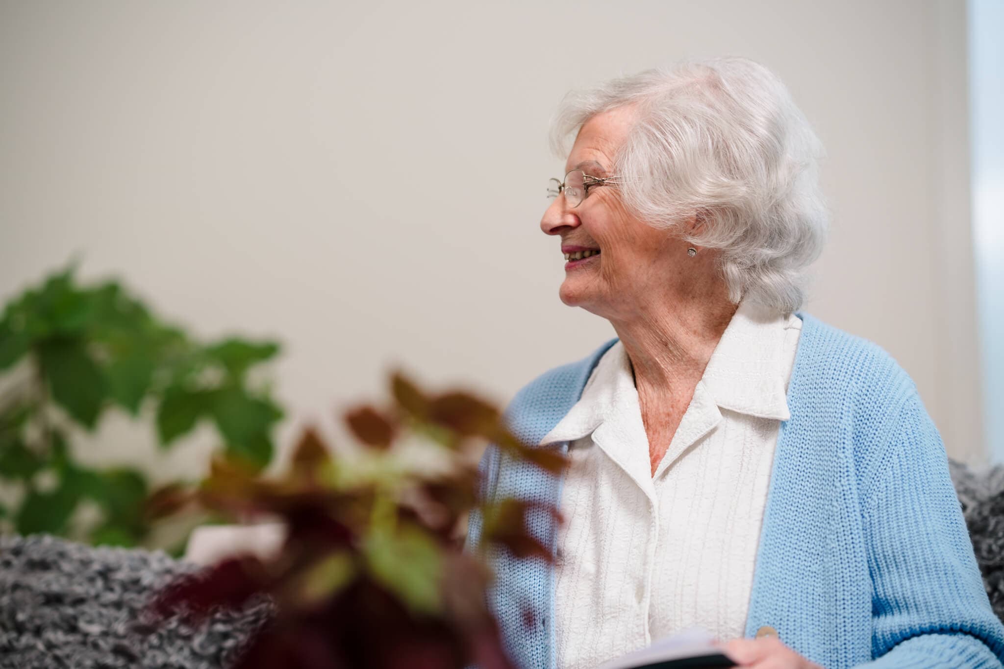 An elderly woman with white hair and glasses, wearing a blue cardigan, smiles while looking to the side. - Home Instead