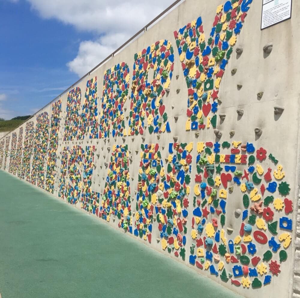 Colorful climbing wall with handholds spelling out "Barry Island" on a sunny day. Green mat at the base and blue sky above. - Home Instead
