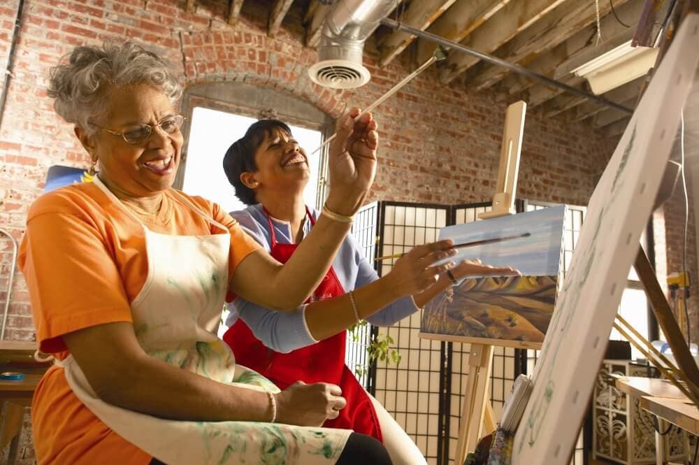 Home Instead Basingstoke picture of two ladies enjoying painting on canvases in a brick-walled art studio with natural light coming through a window