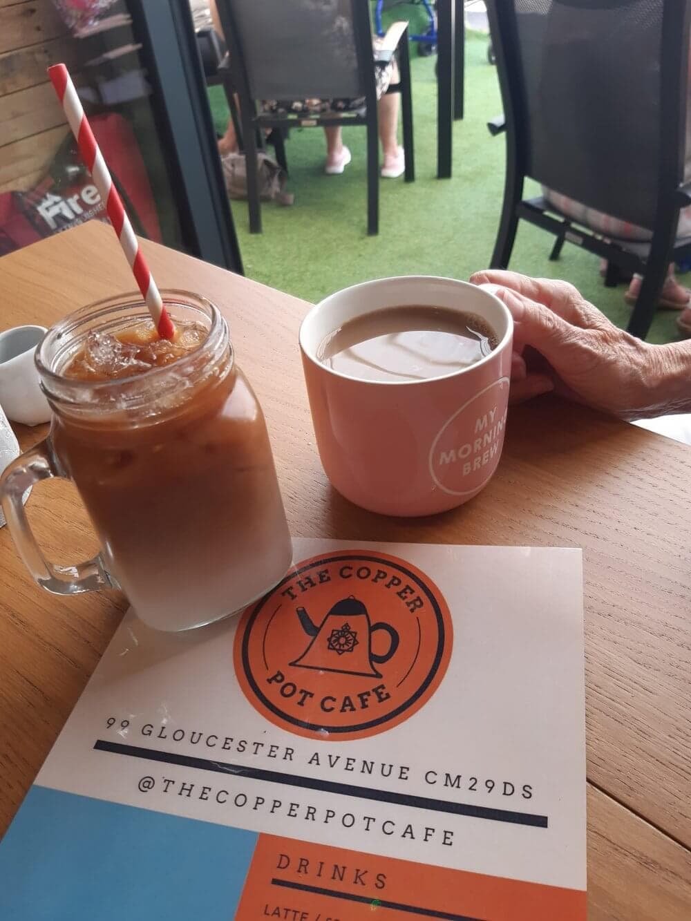 Iced coffee and hot drink on a cafe table with a menu. A hand holds the pink cup. Social media handle is visible. - Home Instead