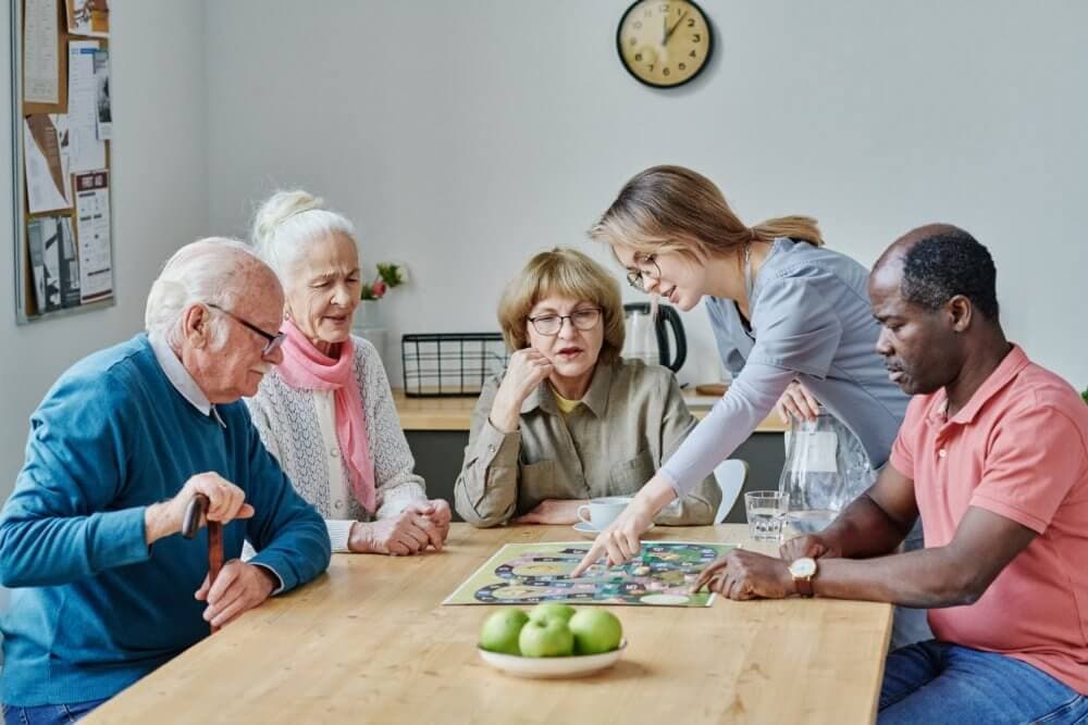 A diverse group of seniors and a young caregiver playing a board game around a table with green apples. - Home Instead