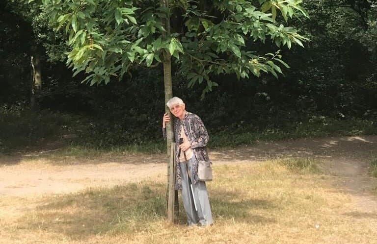 An elderly person, standing outdoors, hugs a tree on a sunny day, surrounded by greenery and a dirt path. - Home Instead