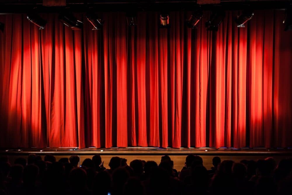 A dimly lit stage with closed red curtains and an audience seated in front, waiting for a performance to begin. - Home Instead