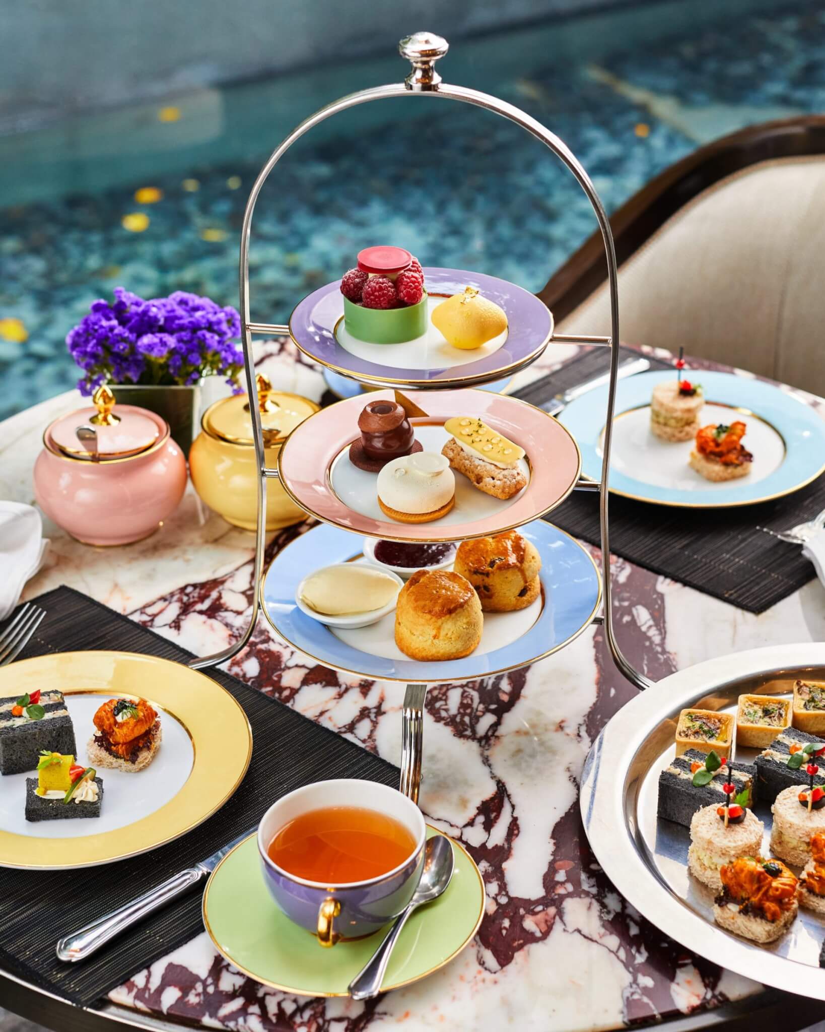 Three-tiered tea stand with assorted pastries, scones, and sandwiches, set on a marble table with a tea cup and flowers. - Home Instead