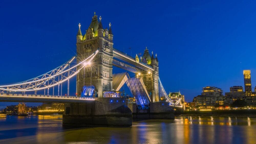 Tower Bridge illuminated at night, with the drawbridge partially raised and city lights reflecting on the river. - Home Instead