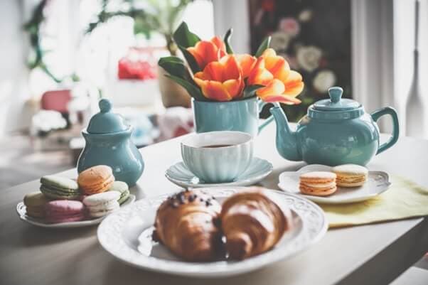 A breakfast table with croissants, macarons, a cup of tea, a teapot, and flowers in a vase. - Home Instead