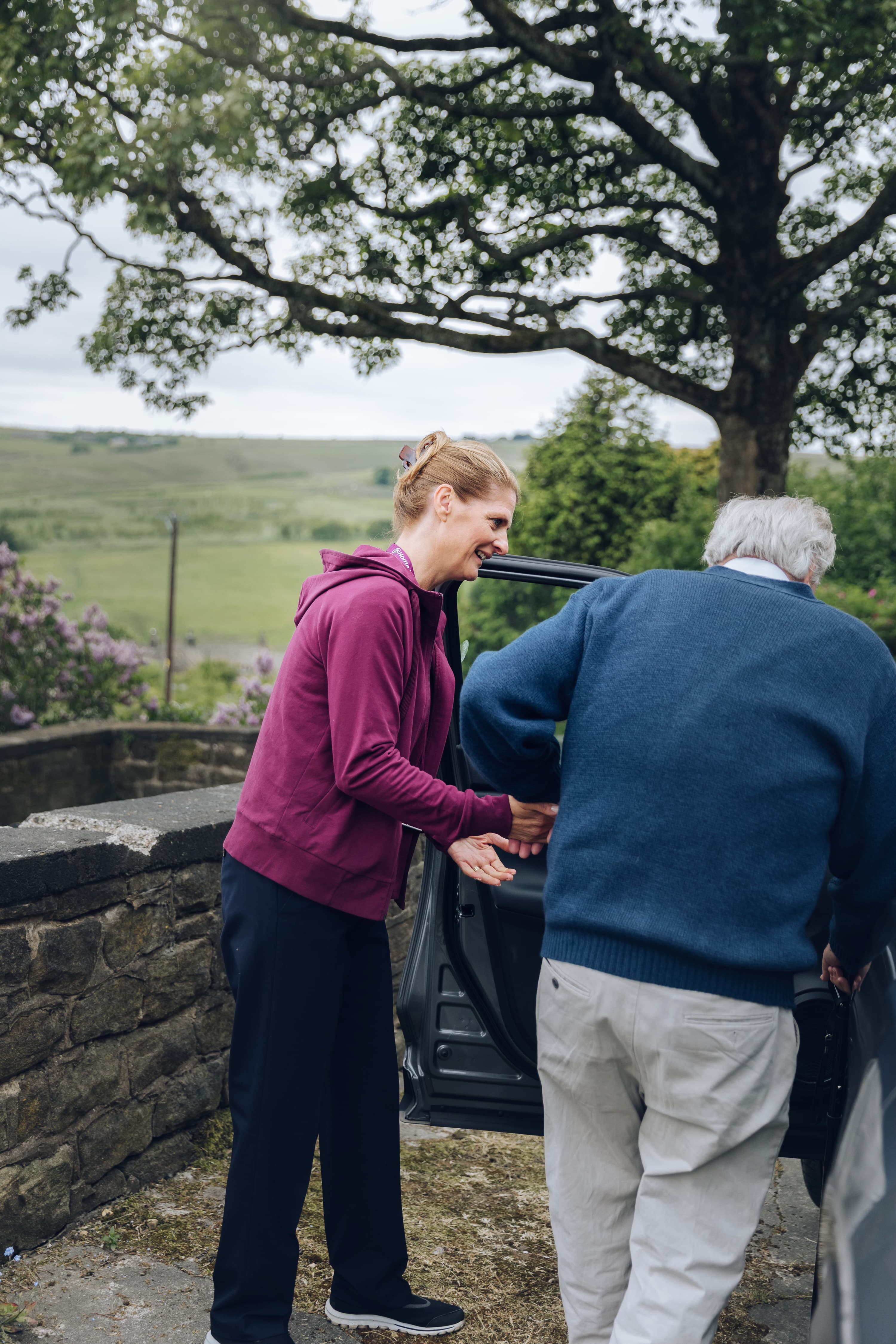 Person in a magenta hoodie assists elderly person into a car against a rural backdrop with trees and stone wall. - Home Instead