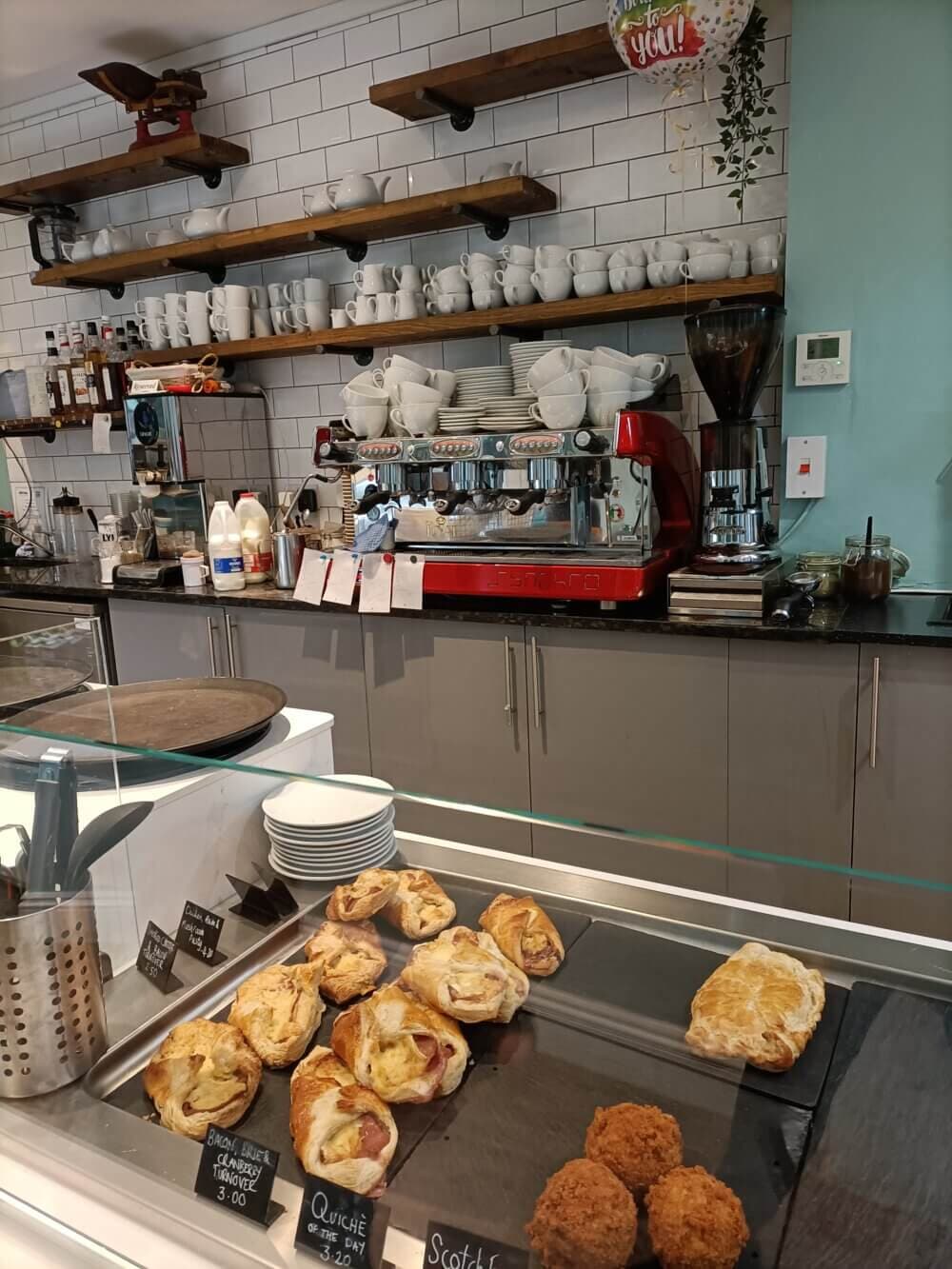 A bakery counter displaying pastries and coffee equipment, with shelves of cups and bottles in the background. - Home Instead