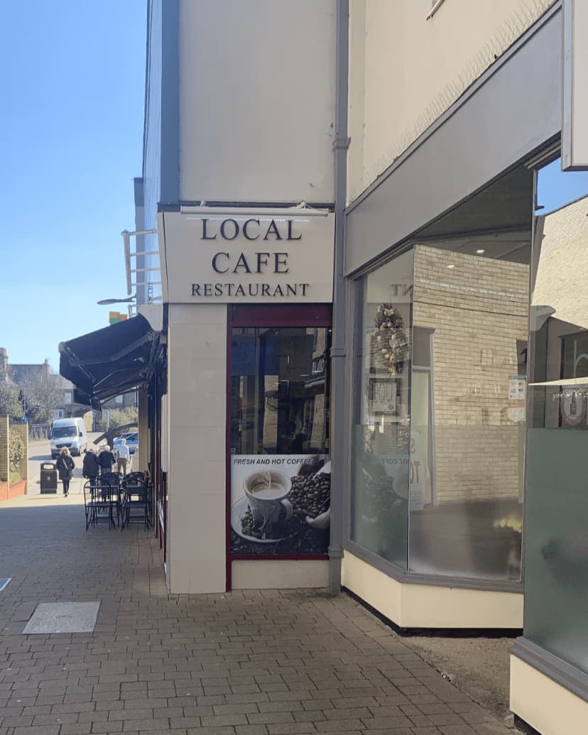 Narrow street with a "Local Cafe Restaurant" sign, an outdoor seating area, and a large window with posters. - Home Instead