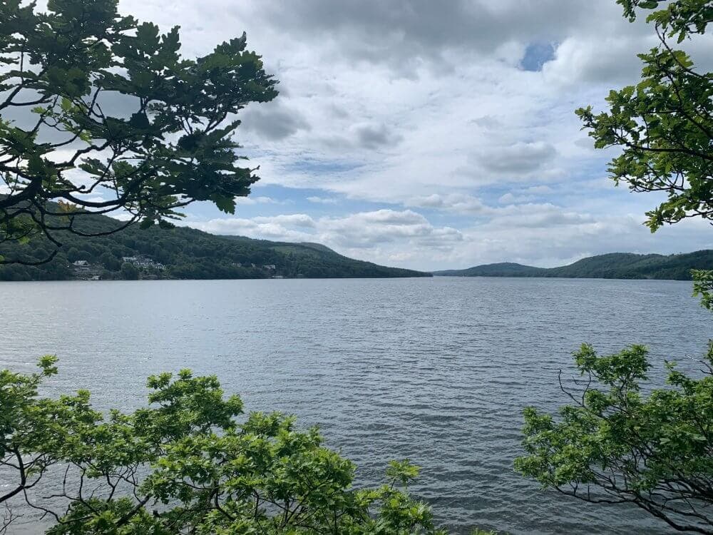 A tranquil lake surrounded by green hills and trees under a partly cloudy sky. - Home Instead