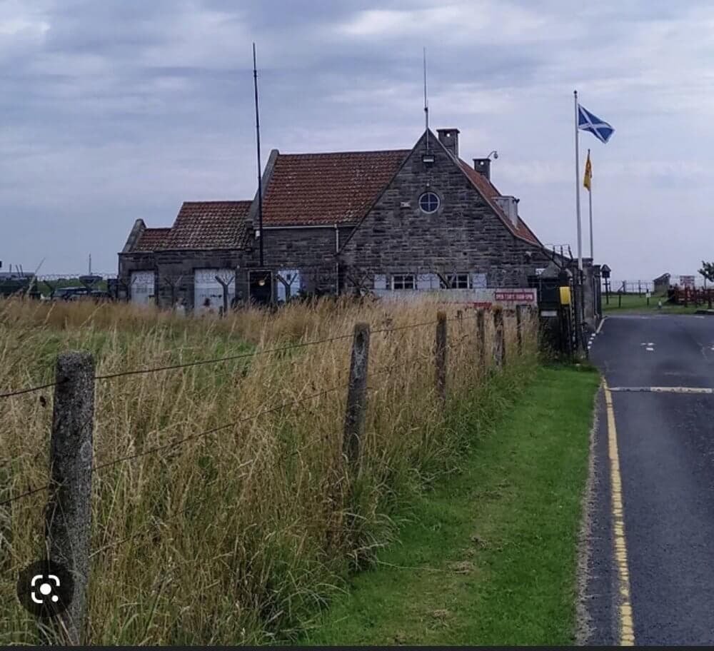 Stone building with red roof, flags flying, and a fence along the pathway on a cloudy day. - Home Instead