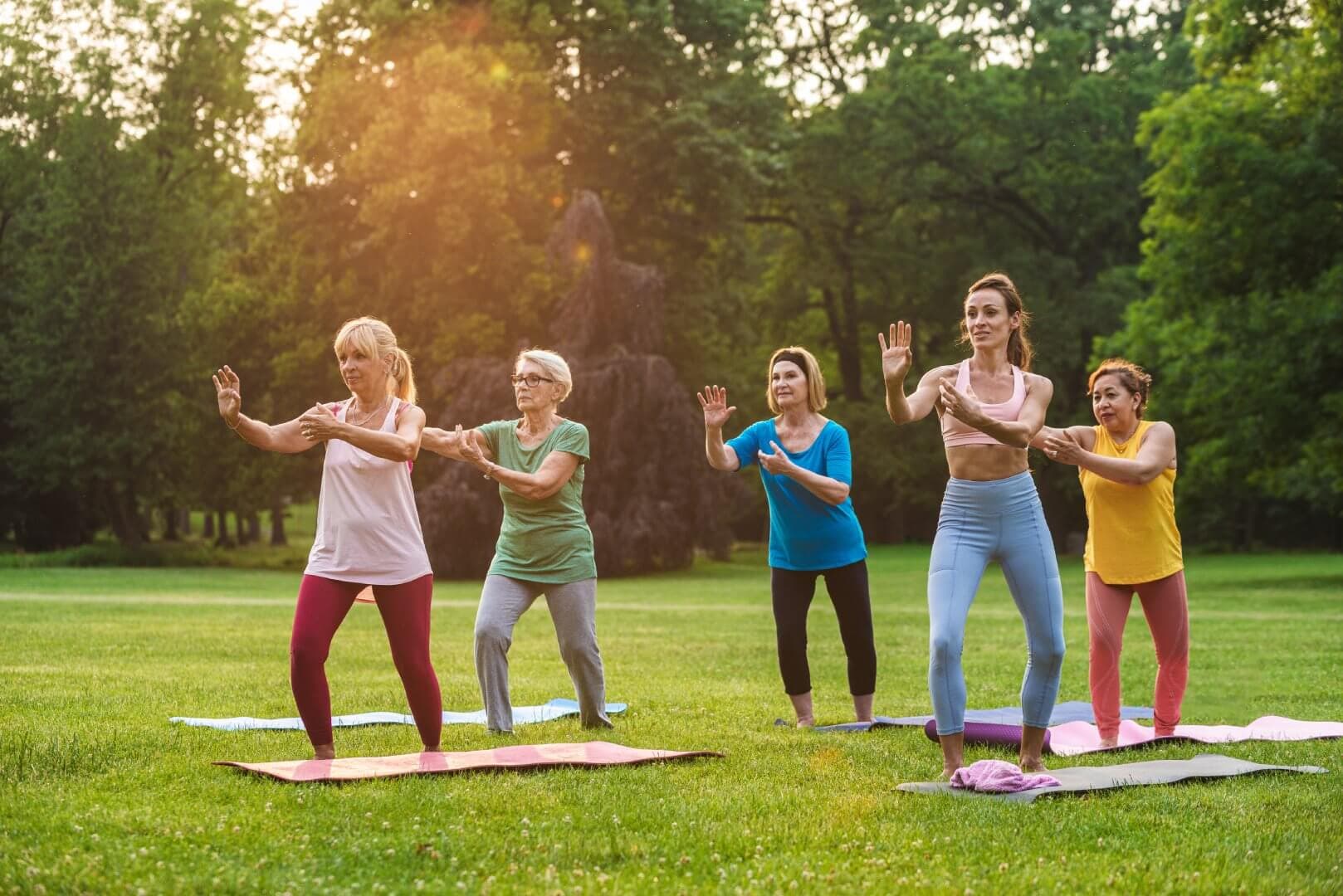 A group of women practicing tai chi on yoga mats in a park. Trees and a fountain are visible in the background. - Home Instead
