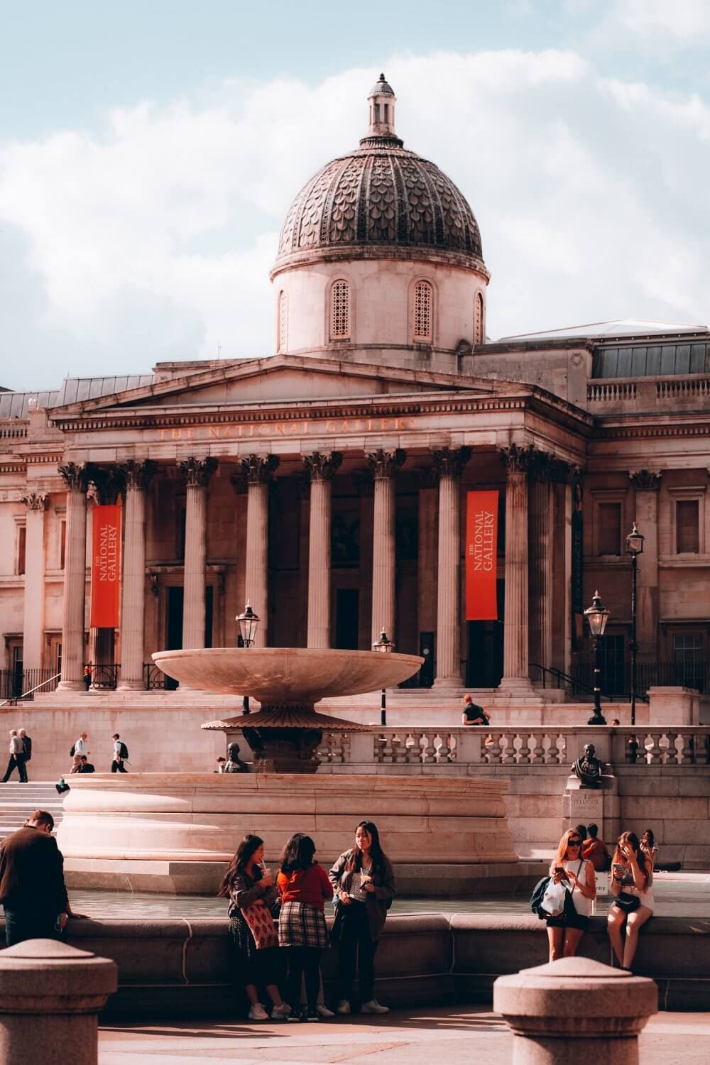 People gather near a fountain in front of a grand, domed building with columns and banners on a sunny day. - Home Instead