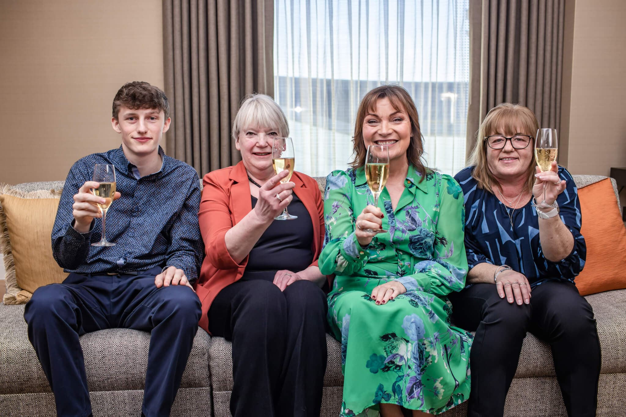 Four people sitting on a couch, smiling and holding glasses of champagne in a warmly lit room with curtains. - Home Instead