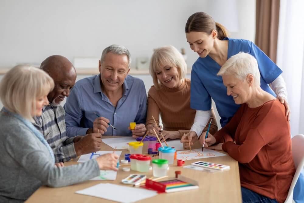 Seniors and a caregiver enjoying an art class, painting and smiling around a table with colorful supplies. - Home Instead