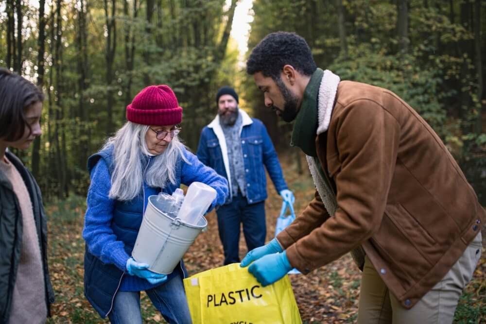 A group of people collects plastic waste in a forest, placing items into a yellow "PLASTIC" bag. - Home Instead