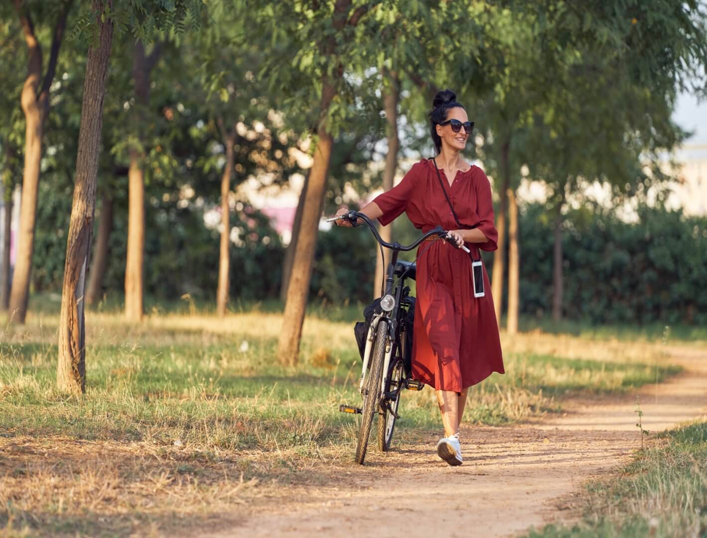 Woman in a red dress walking a bicycle along a tree-lined path, smiling and wearing sunglasses. - Home Instead