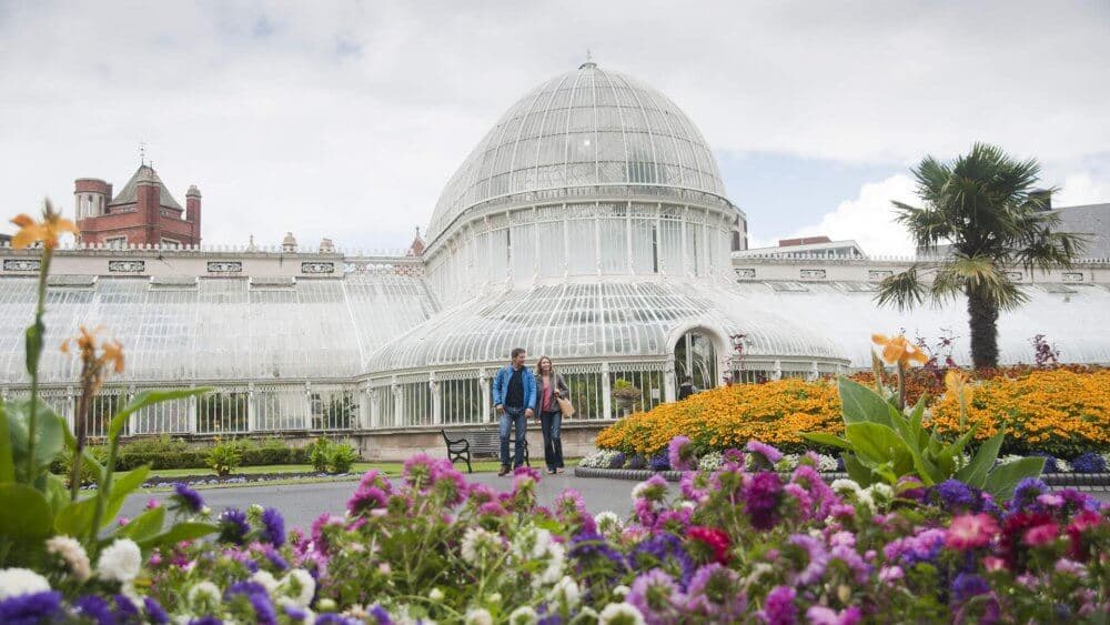 Two people walk by the glass-domed Palm House among colorful flowers at a botanical garden on a partly cloudy day. - Home Instead