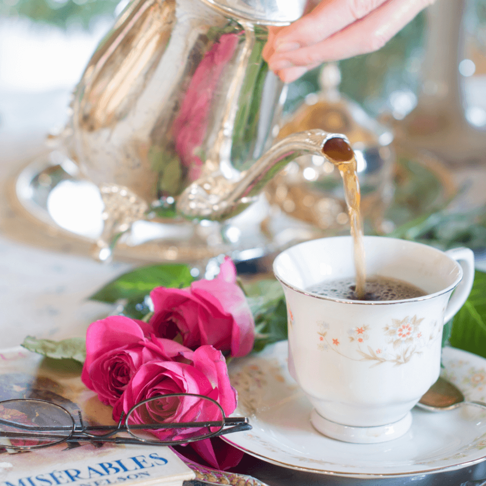 A hand pours tea from a silver teapot into a floral teacup, with pink roses, eyeglasses, and a book nearby. - Home Instead