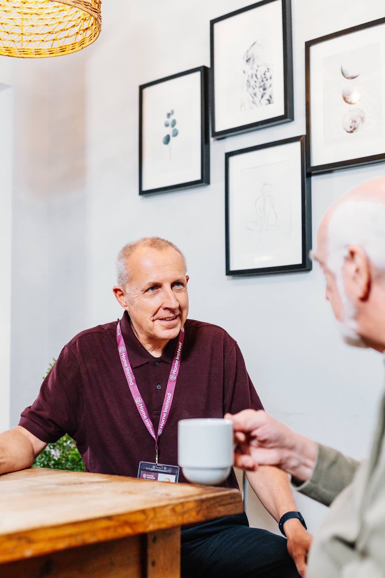 Two men talking at a table, one holding a cup. Artworks hang on the wall behind them. - Home Instead