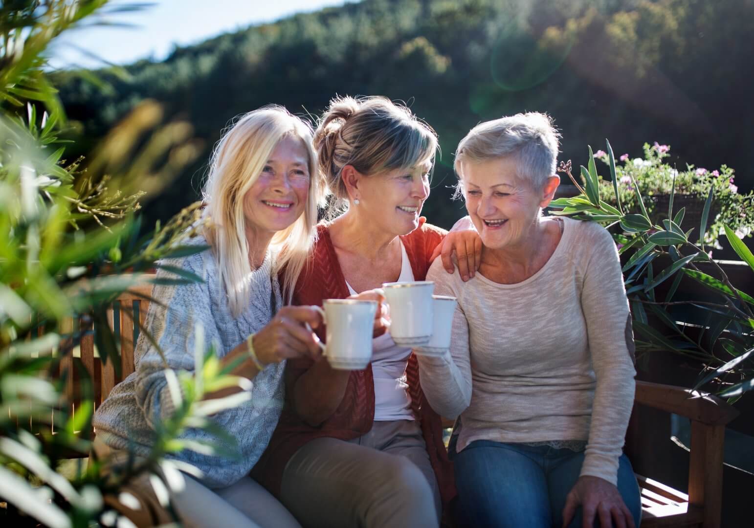 Three elderly women sitting outside, smiling and holding mugs, surrounded by green plants and trees. - Home Instead Poole