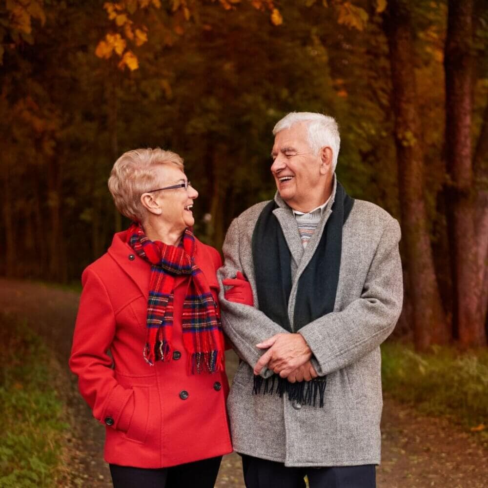 Elderly couple in autumn park, smiling and holding arms, wearing warm coats and scarves. - Home Instead