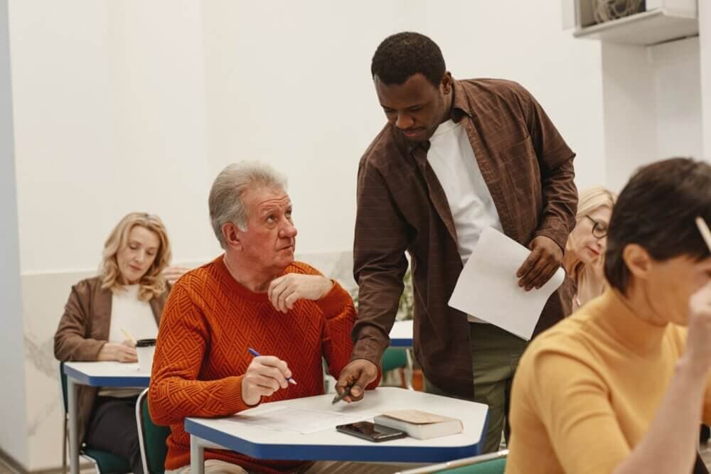 An instructor assists an elderly student in an orange sweater during a classroom activity, while others work. - Home Instead