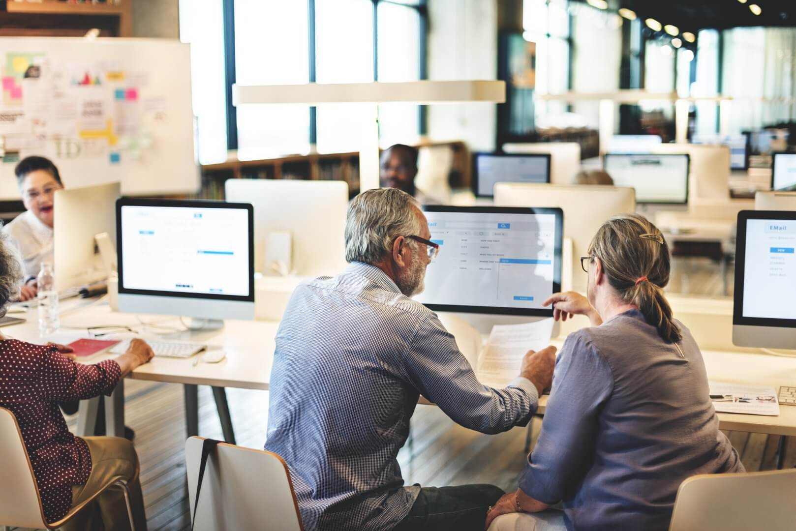 Two people are working together on a project at a computer in a busy office with others working in the background. - Home Instead