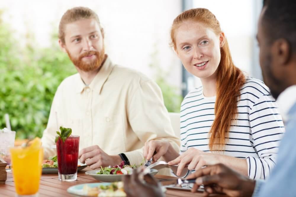 Three people enjoying a meal together at an outdoor table, engaged in conversation and dining. - Home Instead