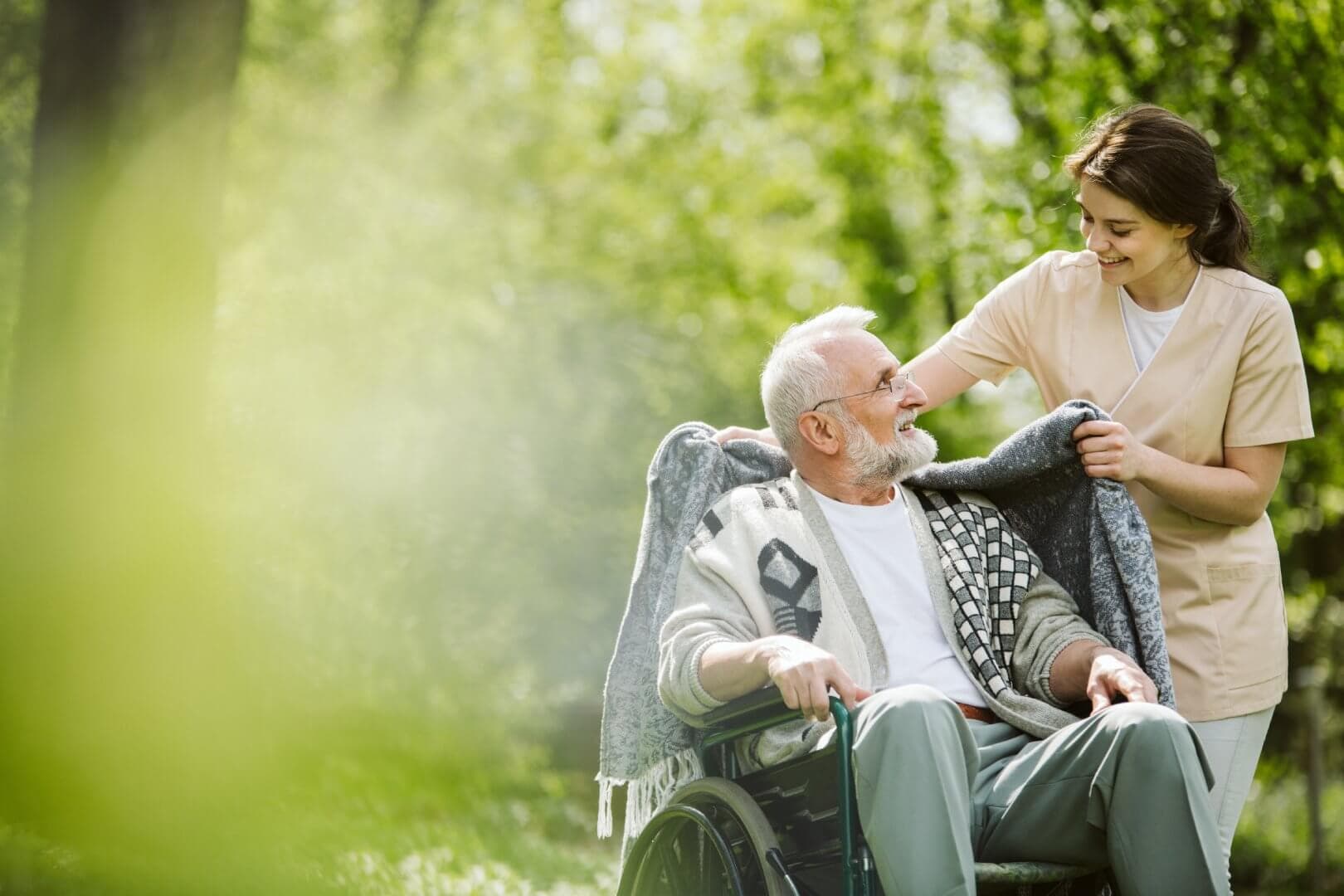 A caregiver wraps a blanket around a smiling elderly man in a wheelchair outdoors on a sunny day. - Home Instead