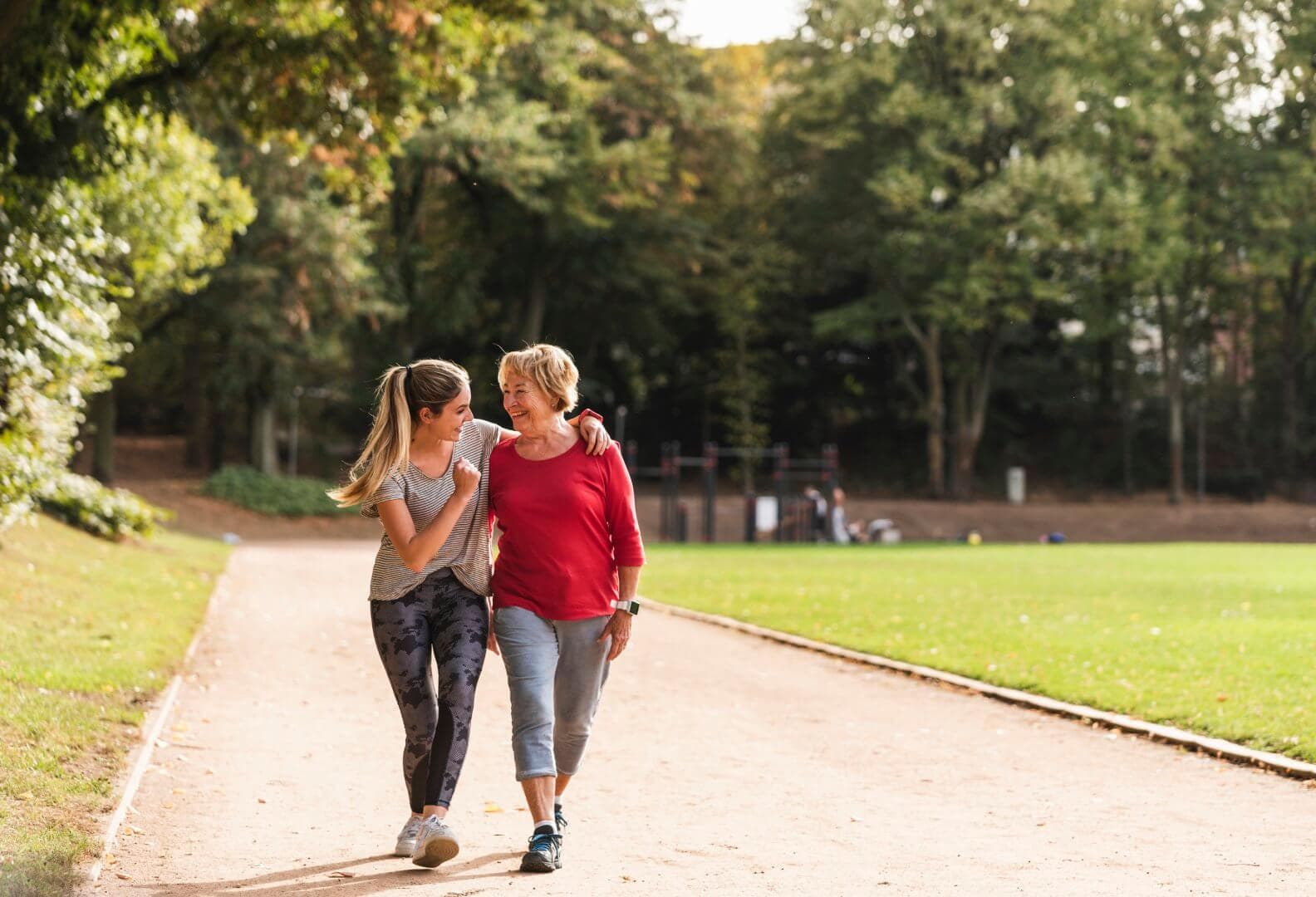 Two women walking and smiling on a path in a park, with one arm around each other. Trees and greenery in the background. - Home Instead
