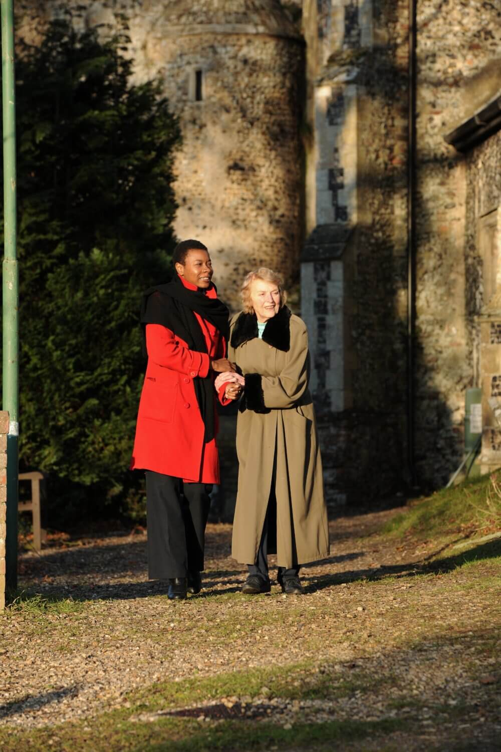 Two women walking arm-in-arm outside a stone building, both wearing long coats, with sunlight casting shadows. - Home Instead