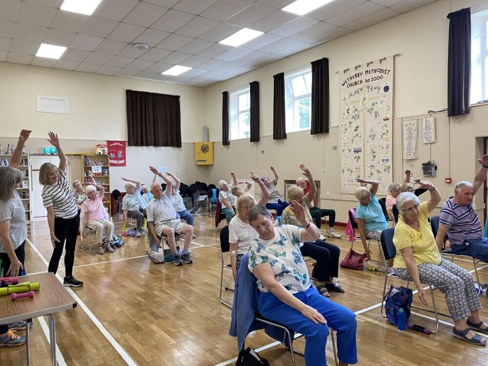 A group of seniors participate in a seated exercise class in a community hall, raising one arm in unison. - Home Instead