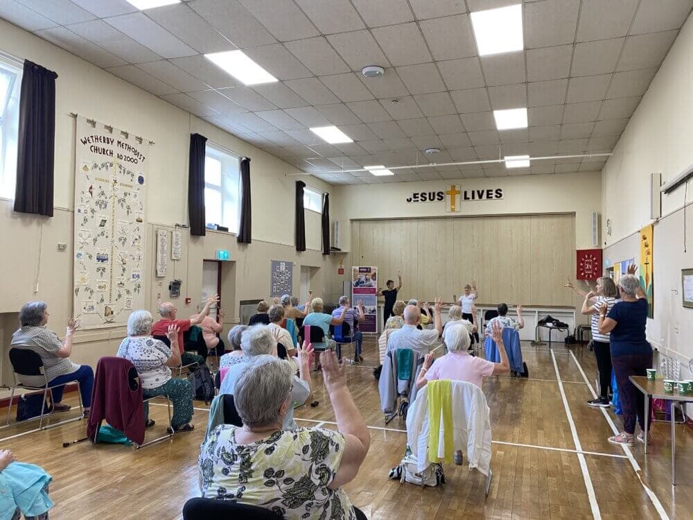 A group of elderly people participate in a seated exercise class in a room with a cross and "Jesus Lives" on the wall. - Home Instead