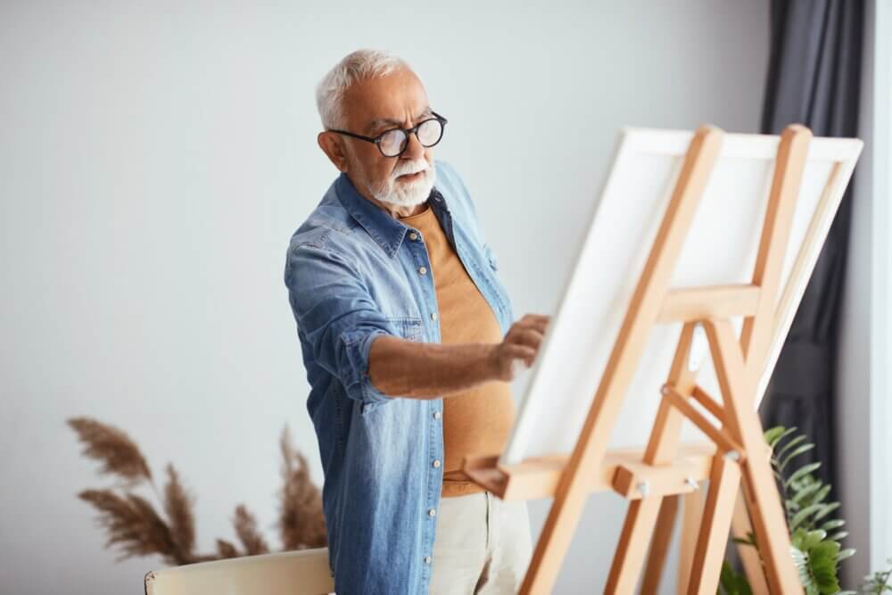 Elderly man with glasses painting on a canvas at an easel in a bright room. - Home Instead
