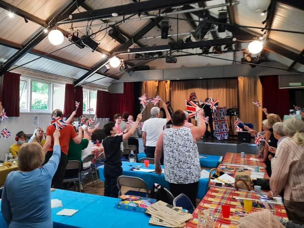 People waving Union Jack flags inside a hall, decorated with flags and tables, celebrating an event. - Home Instead