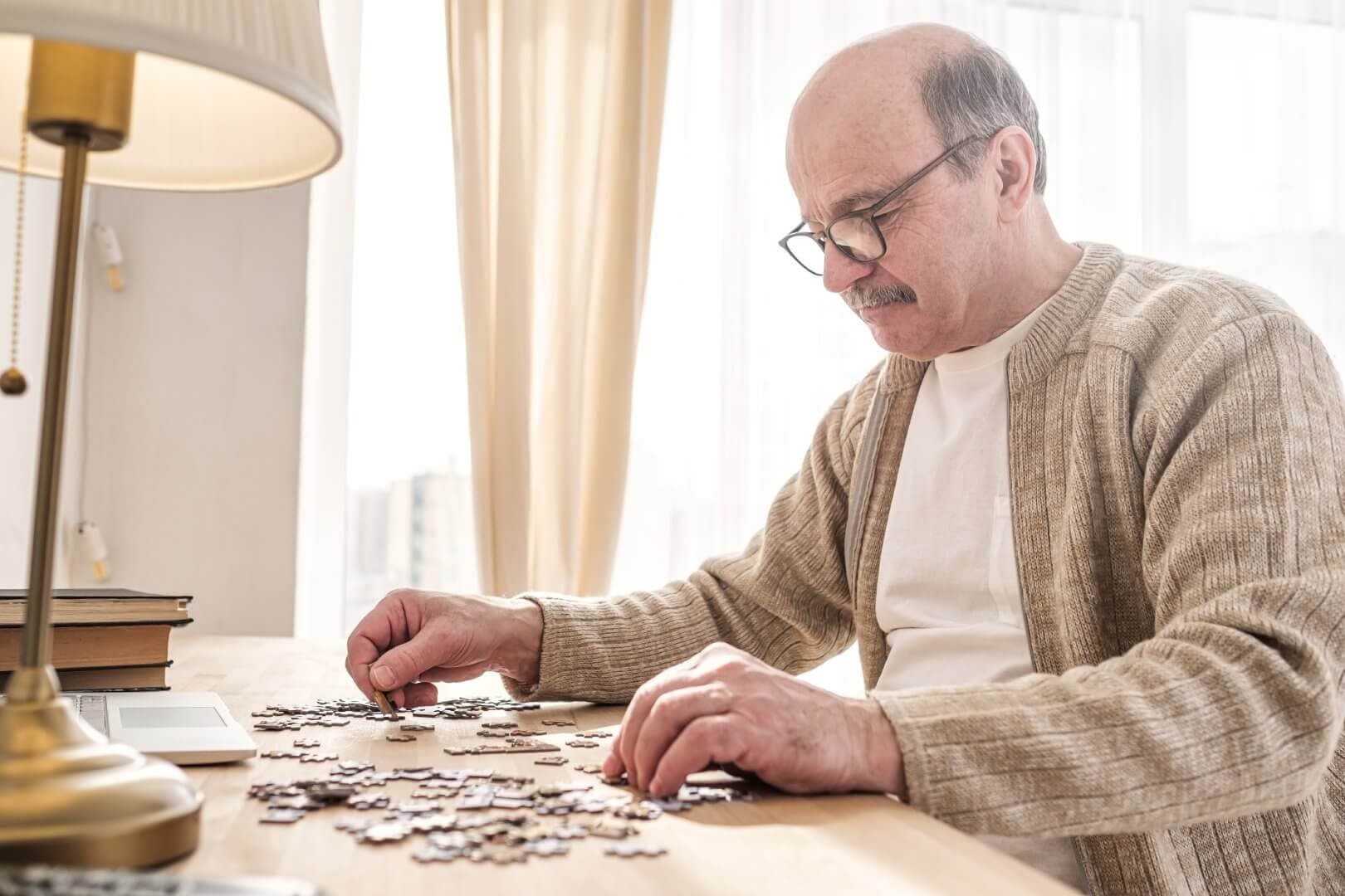 Elderly man with glasses focuses on assembling a jigsaw puzzle at a bright, sunlit table beside a lamp and books. - Home Instead