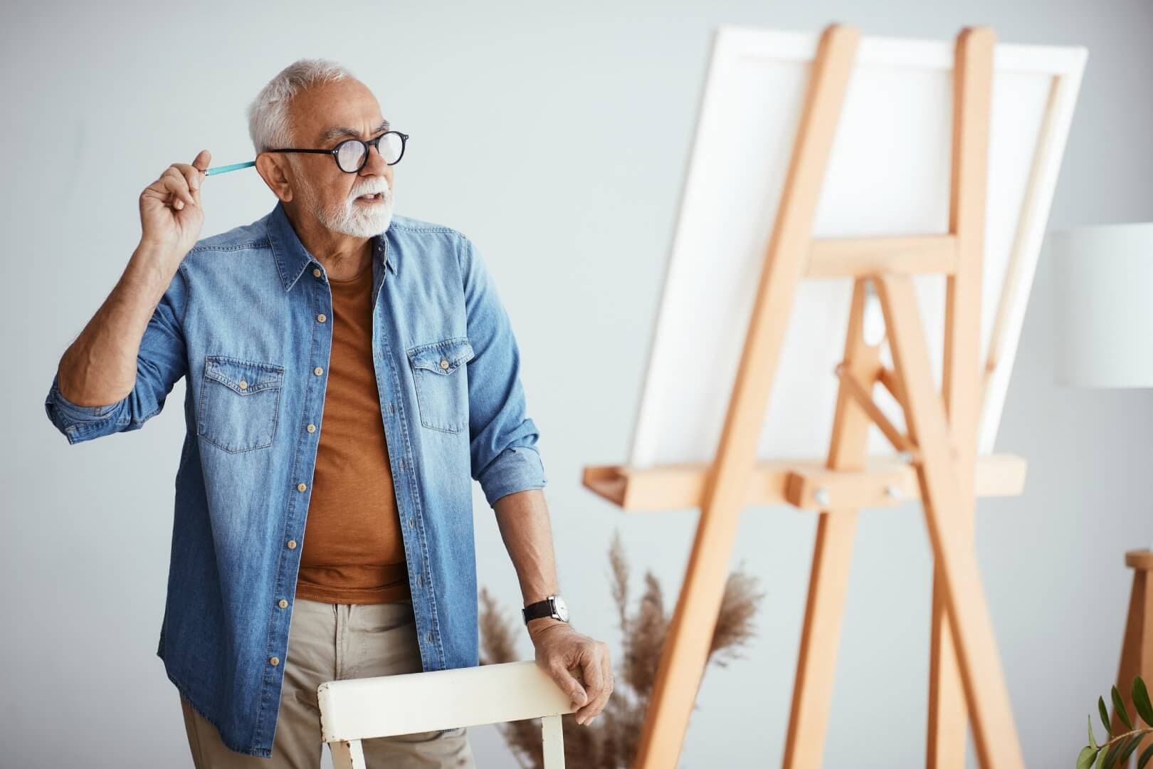 A man wearing glasses and a denim shirt holds a paintbrush, standing in front of an easel with a blank canvas. - Home Instead