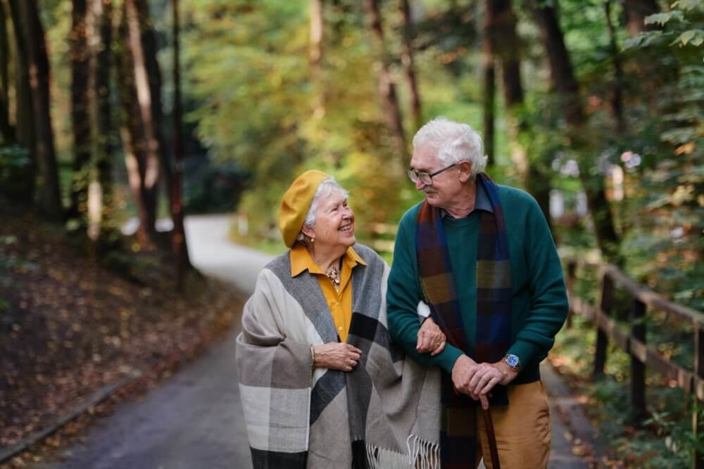 Elderly couple walking arm in arm down a forest path, wearing scarves and smiling at each other. - Home Instead