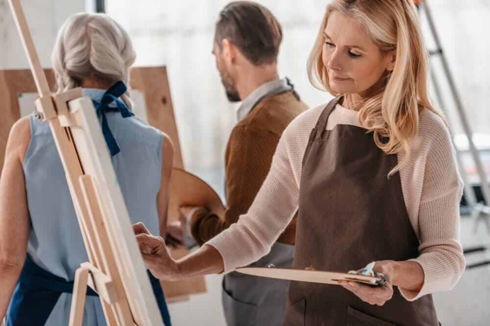 Three adults in an art class, with two women painting on canvases and a man drawing on a board in the background. - Home Instead