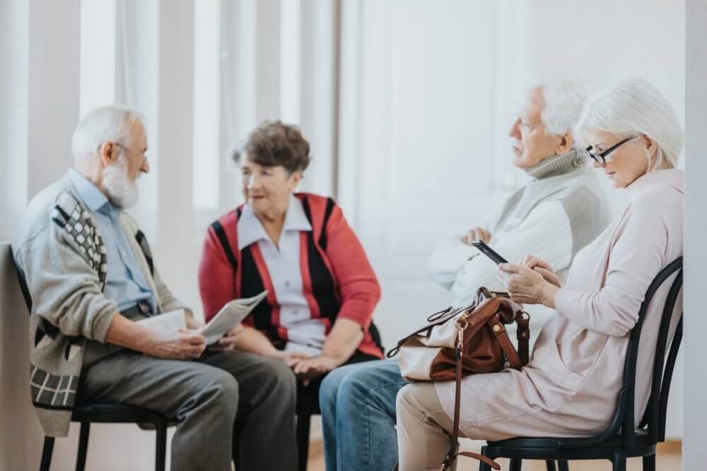 Four older adults sitting on chairs; two chatting, one using a smartphone, and one reading a newspaper. - Home Instead