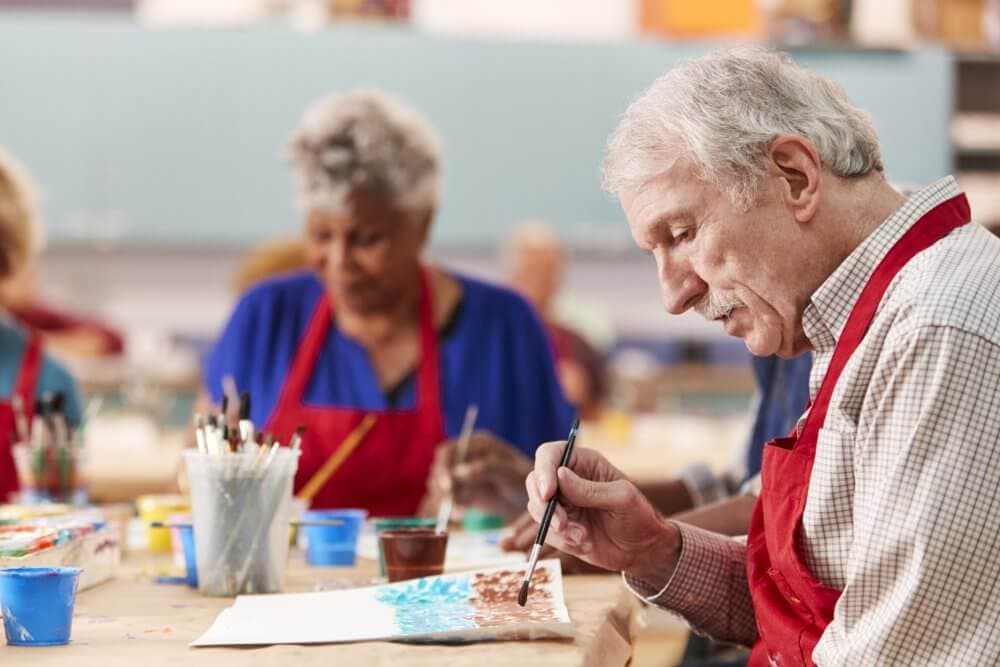 Seniors in a painting class, focused on their artworks, with paint supplies and brushes on the table. - Home Instead