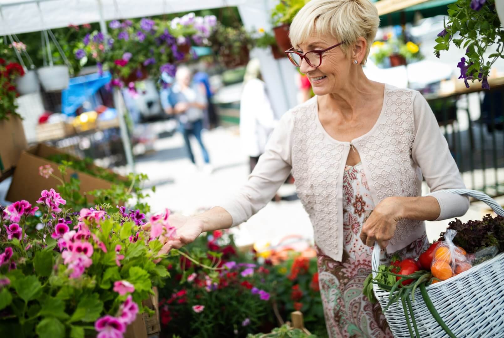 A woman with short hair and glasses holds a basket while selecting flowers at an outdoor market. - Home Instead