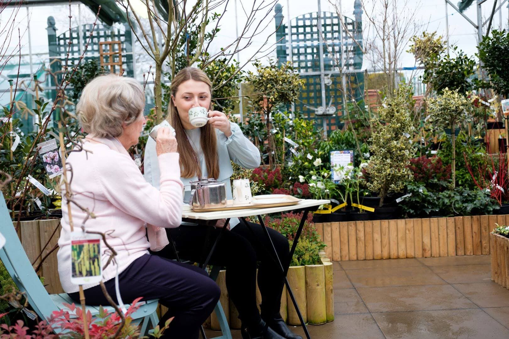 Two women sitting at an outdoor café table, drinking tea with plants and greenery surrounding them. - Home Instead