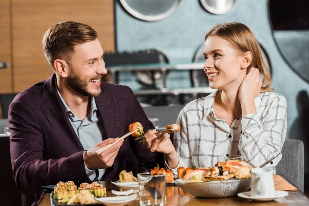 A smiling couple enjoys a sushi meal together at a restaurant, using chopsticks to share food. - Home Instead